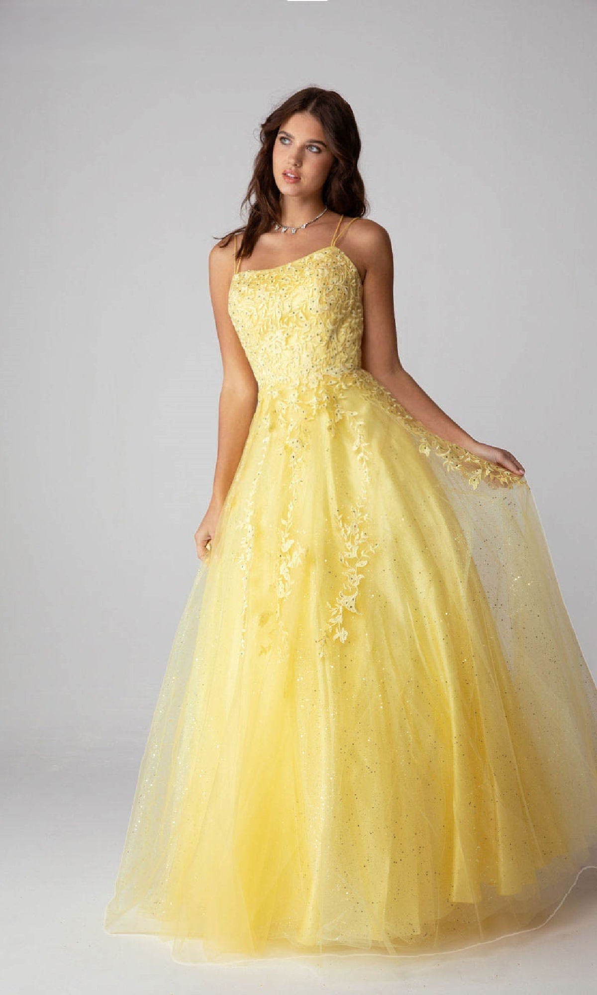 A woman in the Lace-Up Long Lace Prom Ball Gown: Eureka CC0240, featuring yellow lace, floral embroidery, a tulle skirt, and a stylish lace-up back, stands against a plain backdrop.